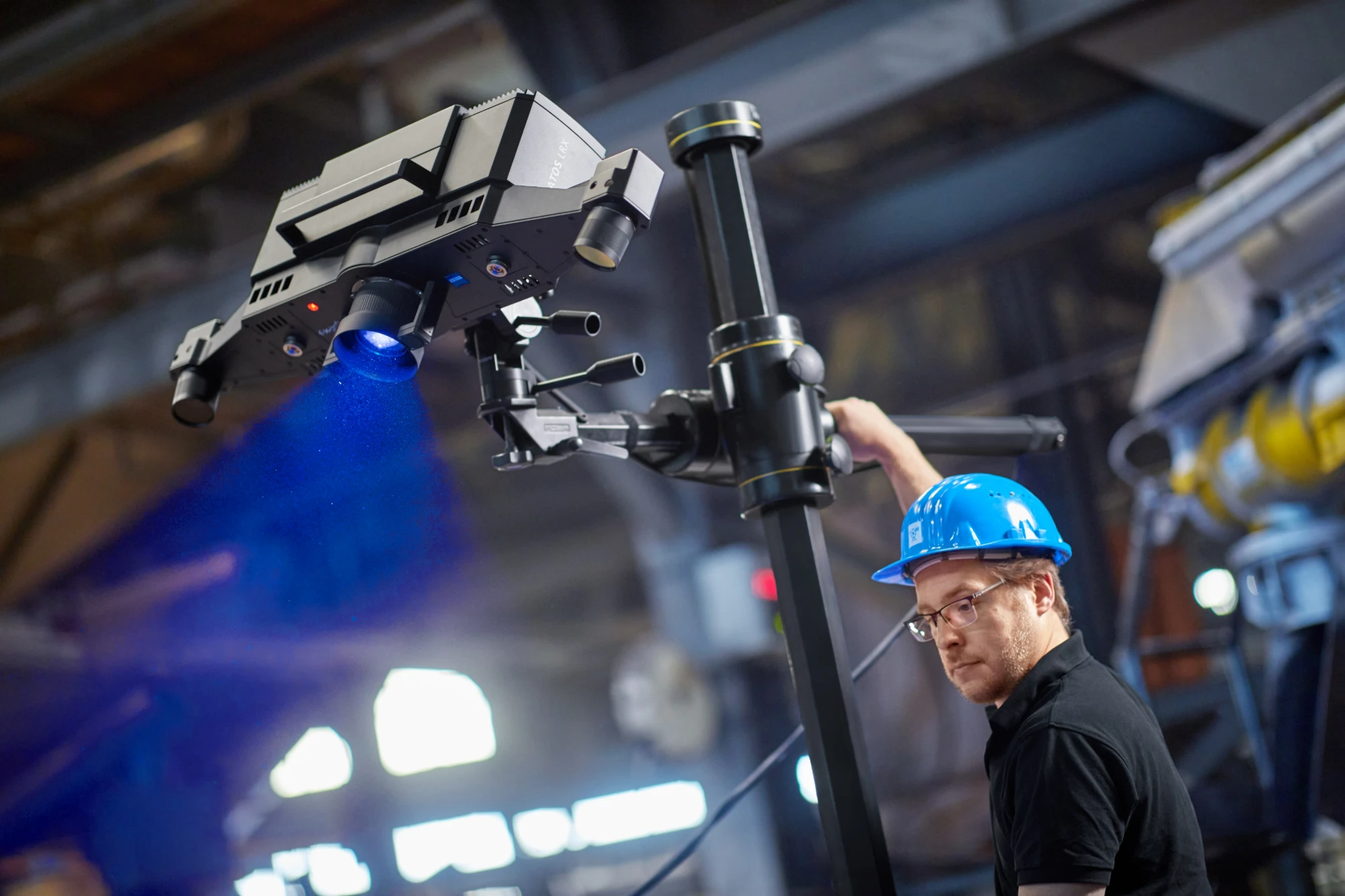 Technician in a blue hard hat aiming a ZEISS ATOS LRX scanner, its blue measuring light falling across a heavy production hall