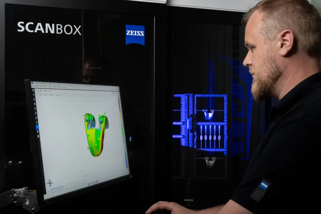 Engineer at a ZEISS ScanBox reading a colour deviation map on screen while the cell scans a part behind the glass
