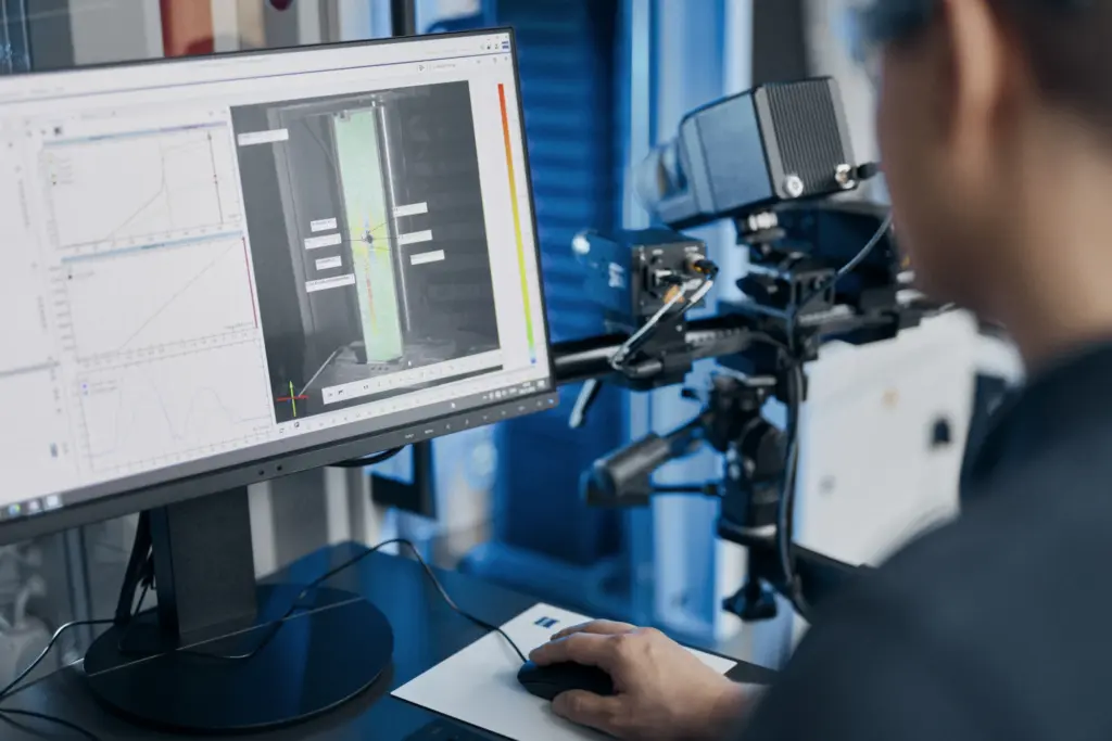 Engineer at a monitor showing ARAMIS strain measurement of a tensile specimen, the sensor aimed at the test rig