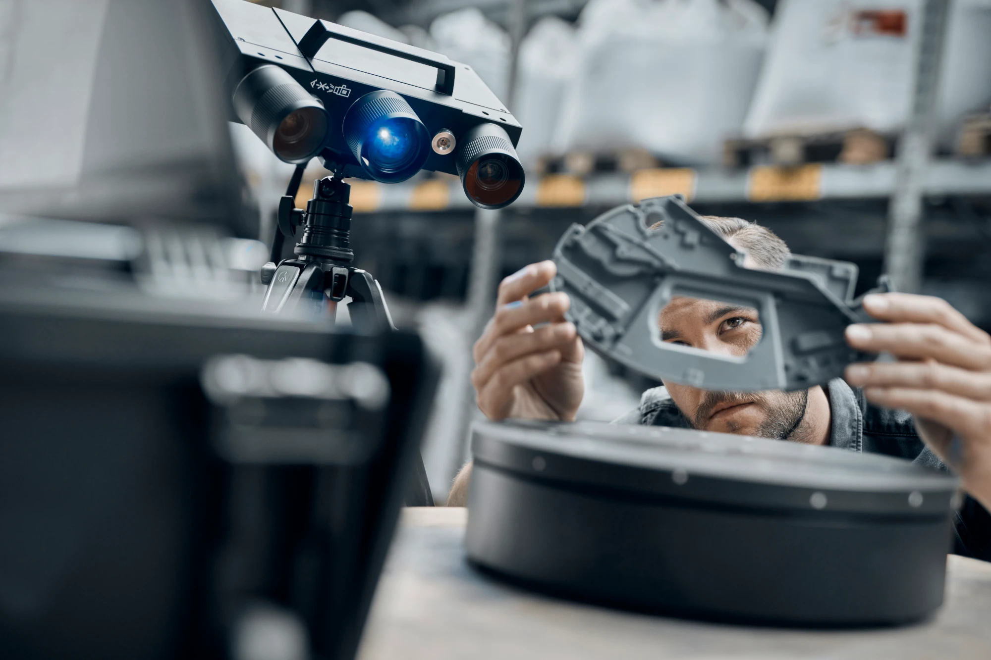 Engineer holding a dark cast part up beside a tripod-mounted ZEISS ATOS Q scanner in a warehouse